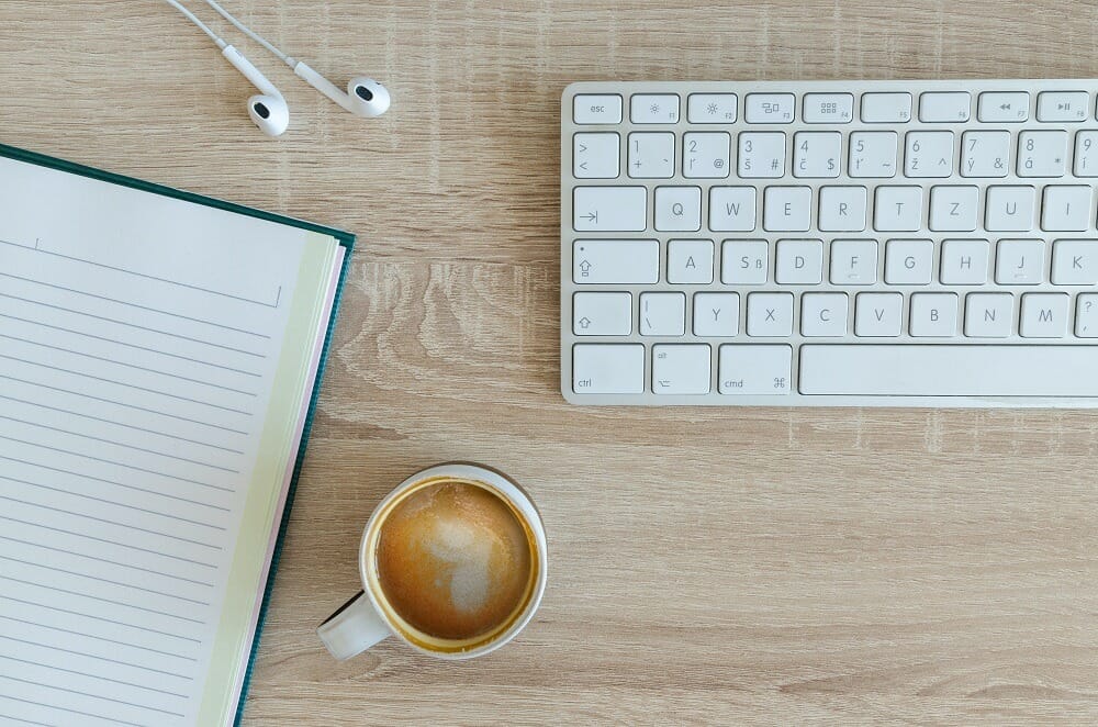 white keyboard, notebook, and coffee cup on a light wooden desk