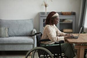 Side view portrait of young African-American woman using wheelchair while working from home in minimal grey interior