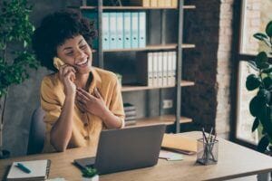 Photo of sincere cheerful mixed-race laughing girl speaking by phone with her friend colleague, enjoying her break with pens notepad on desktop